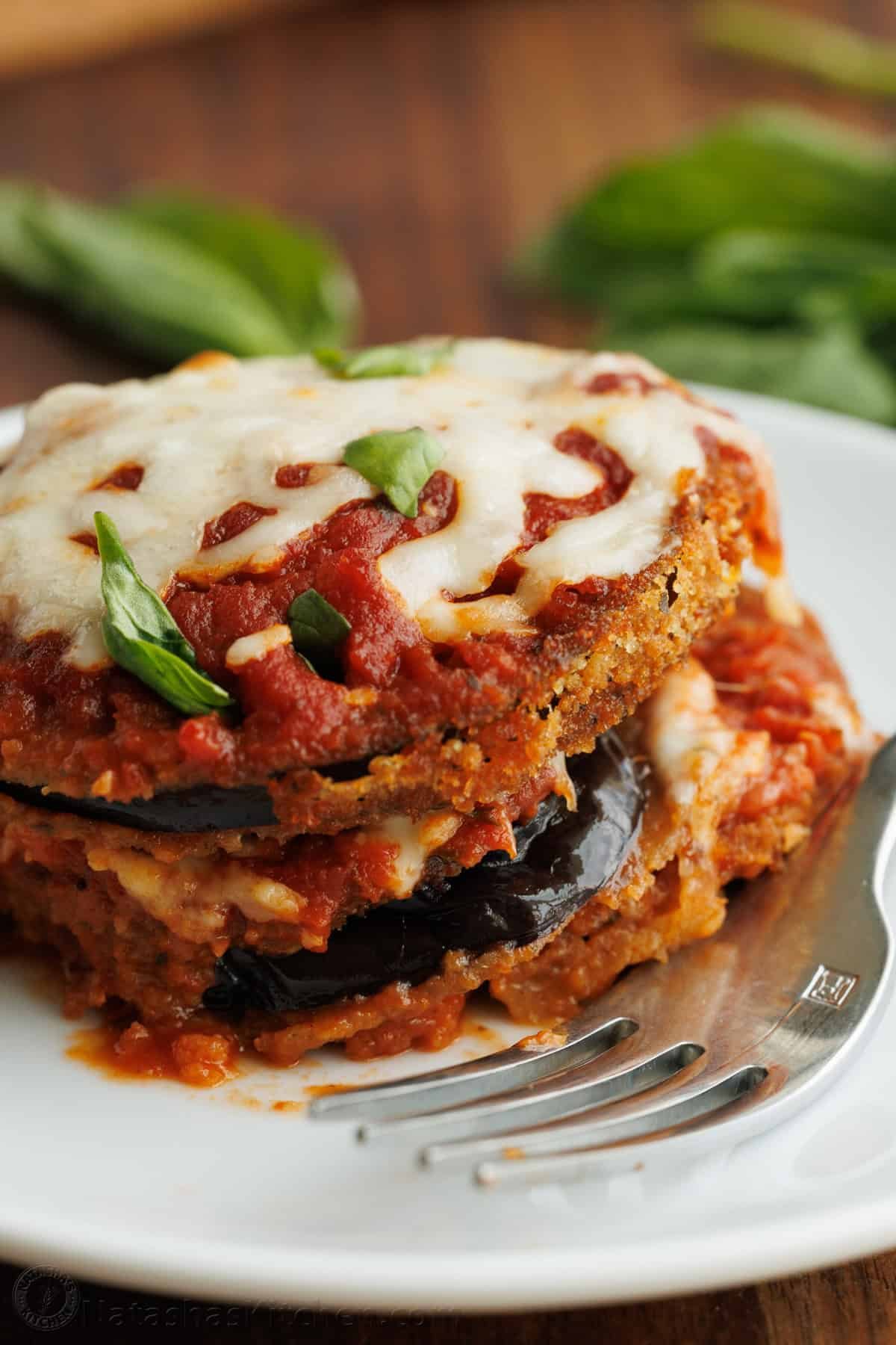 Eggplant Parmesan: Crispy breaded eggplant, layered with marinara sauce and mozzarella cheese, baked to perfection into a saucy and cheesy casserole. Breaded eggplant Parmesan layered with marinara sauce and mozzarella cheese, and garnished with fresh basil served on a plate with a fork