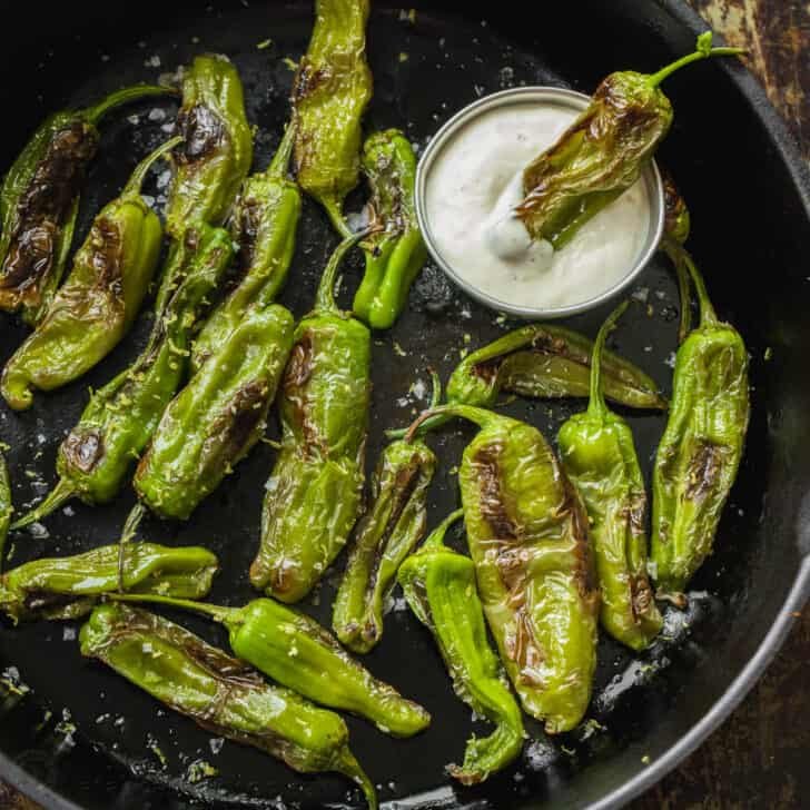 Blistered shishito peppers in skillet with small bowl of aioli