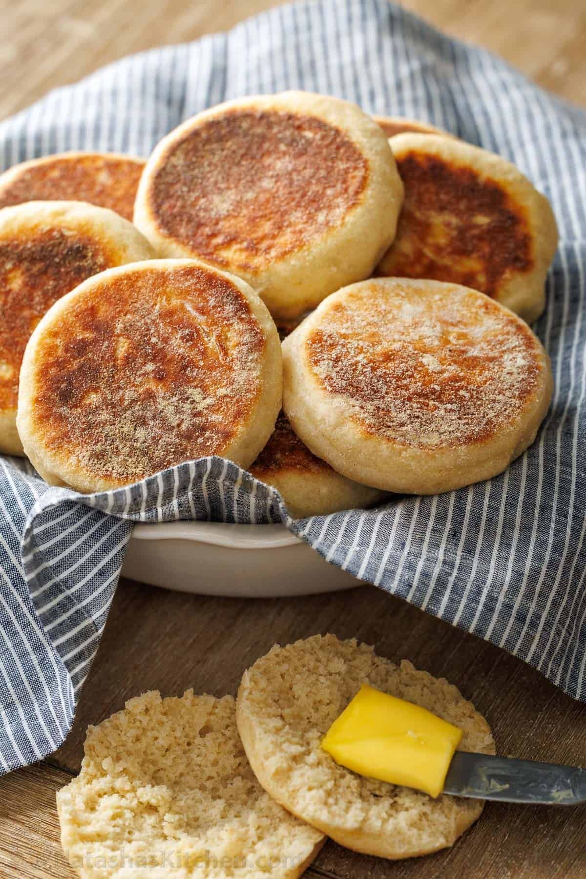 Butter spread on homemade english muffin with bowl of toasted muffins