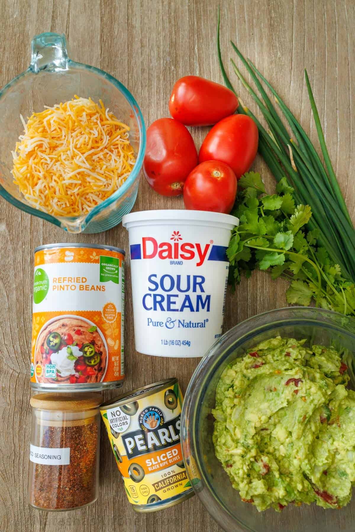 Ingredients for 7 layer dip: refried beans, shredded cheese, sour cream, black olives, guacamole, taco seasoning, cilantro, green onions, Roma tomatoes
