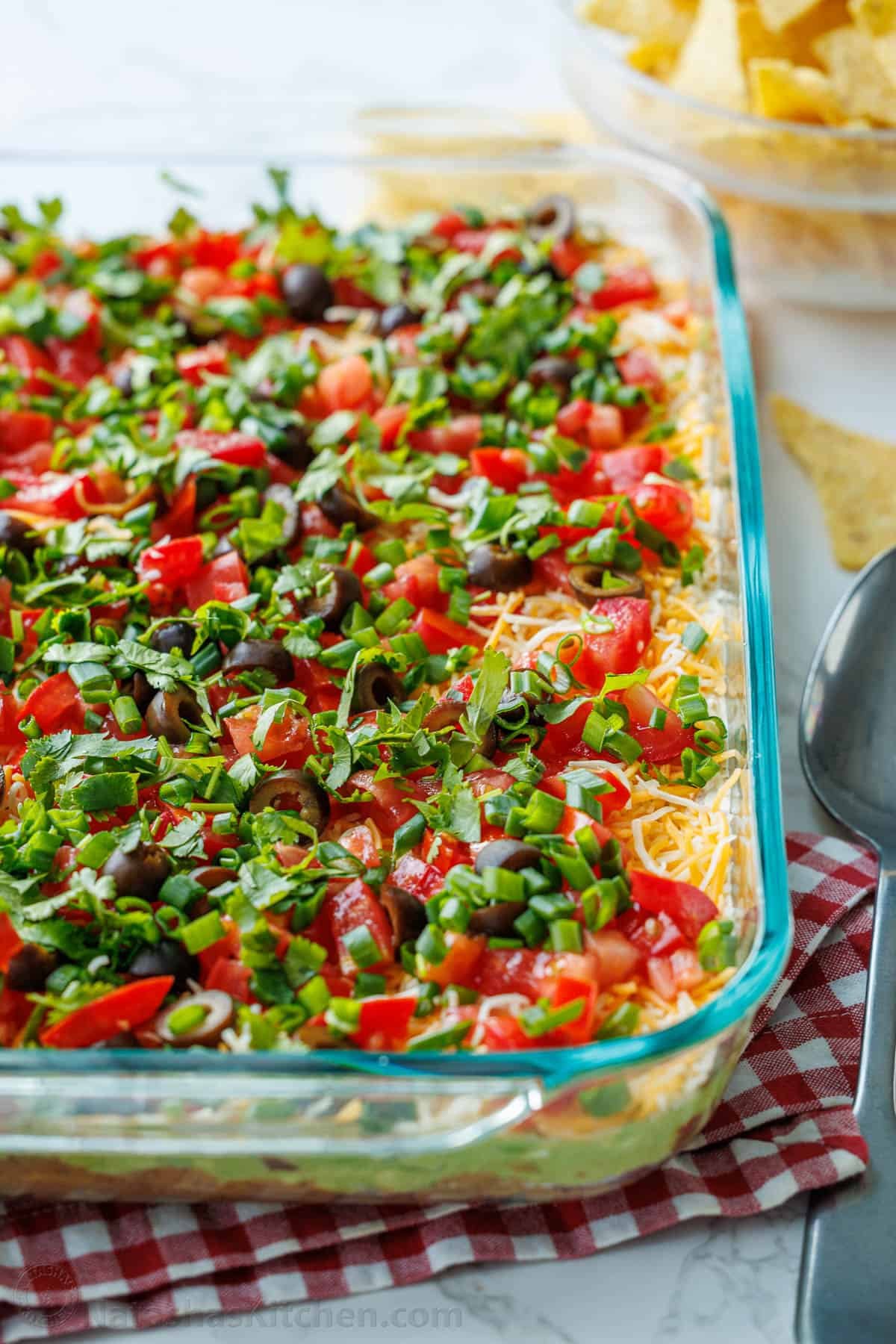 Assembled 7-Layer Dip in a glass pan, showing the top layer of cheese, tomatoes, green onion, and black olives.