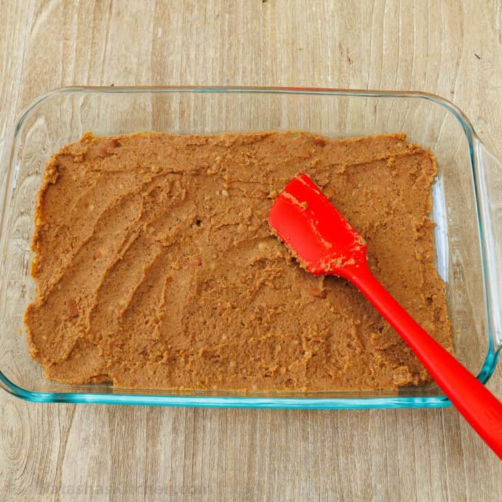 Refried beans mixed with taco seasoning spread into a glass pan for the base layer of 7-Layer Dip