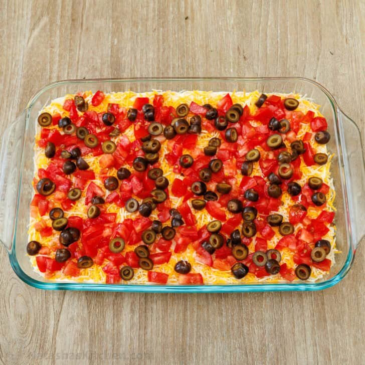 Diced Roma tomatoes and sliced black olives layered on top of the cheese layer in a glass pan for 7-Layer Dip