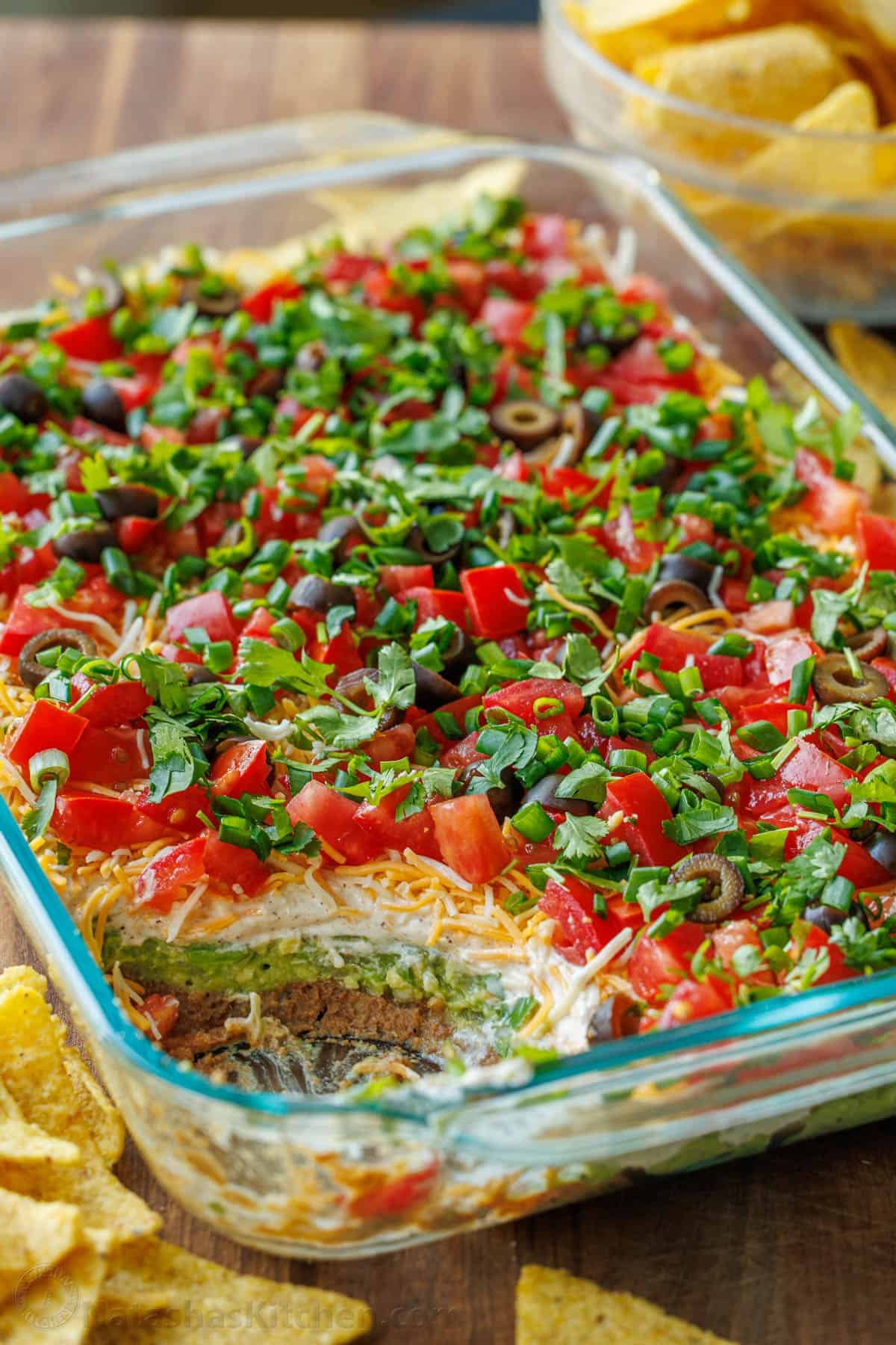 7-Layer Dip in a glass pan, showing each layer, refried beans, sour cream, cheese, tomatoes, green onion, guacamole, black olives with tortilla chips scattered around