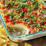 7-Layer Dip in a glass pan, showing each layer, refried beans, sour cream, cheese, tomatoes, green onion, guacamole, black olives with tortilla chips scattered around