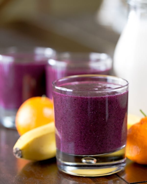 A close up of a glass of a purple smoothie with bananas and mandarins in the background