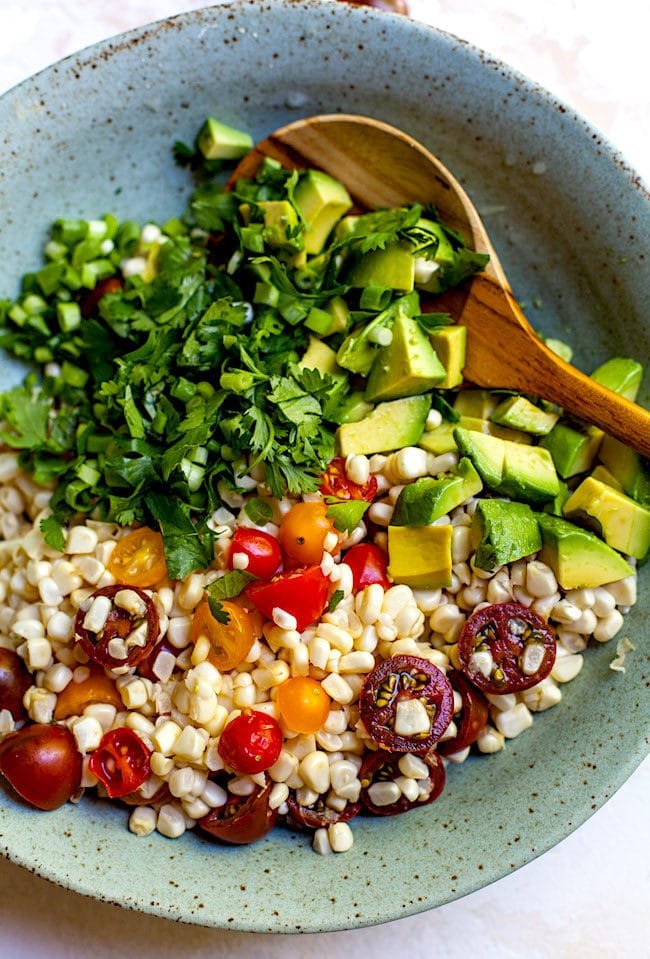 corn, tomatoes, avocado, and cilnatro in bowl with wooden spoon to make corn salad.