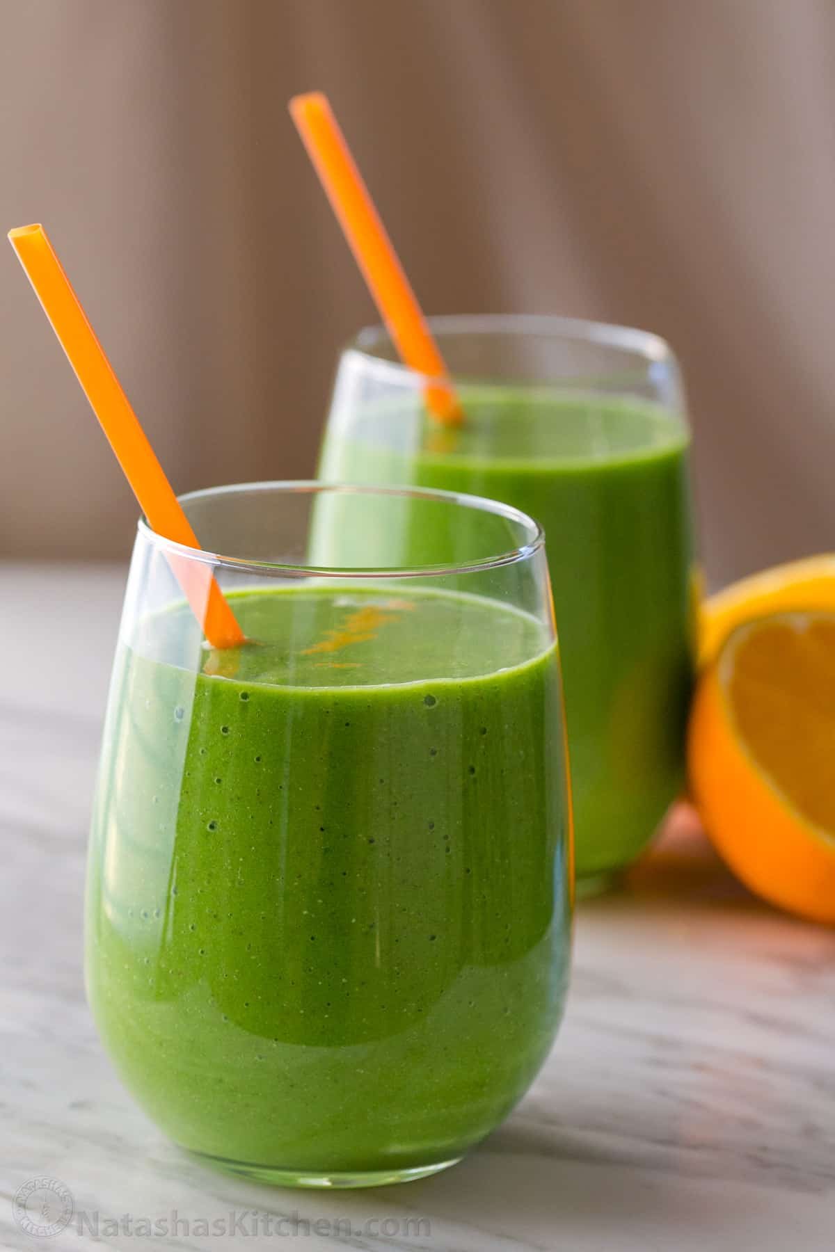 This Green Smoothie recipe is packed with nutritious ingredients like spinach, orange, pineapple, banana, and chia seeds. It's so refreshing. Two green smoothies in glasses with straws. Orange halves resting on the counter next to the glasses.