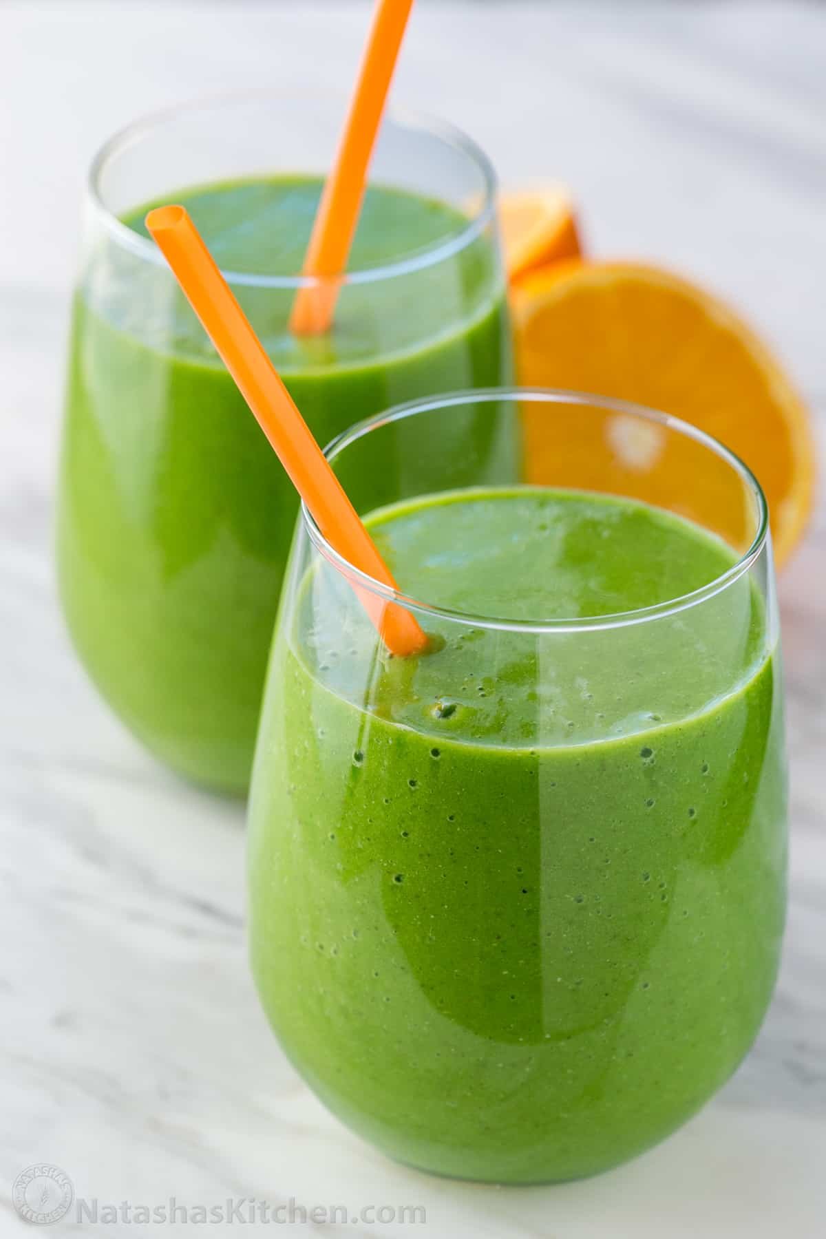 This Green Smoothie recipe is packed with nutritious ingredients like spinach, orange, pineapple, banana, and chia seeds. It's so refreshing. Two green smoothies in glasses with straws. Orange halves resting on the counter next to the glasses.