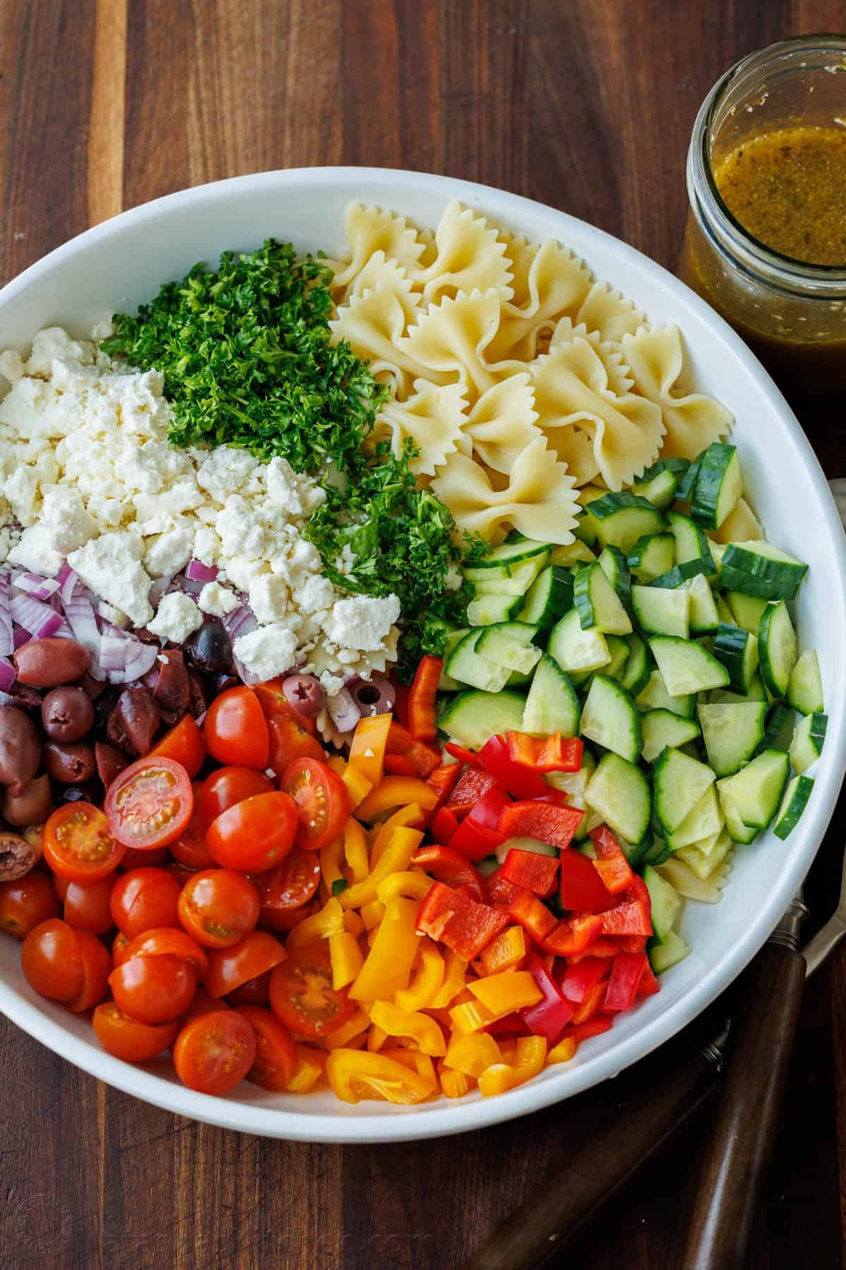 Greek Pasta Salad is a fresh, flavorful, and satisfying dish that brings a burst of Mediterranean flavor to any table. Perfect for any occasion and sure to please. Diced Greek Pasta Salad ingredients in a white serving bowl: bowtie pasta, cucumber, bell pepper, halved cherry tomatoes, Kalamata olives, red onion, feta cheese, and parsley