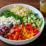 Diced Greek Pasta Salad ingredients in a white serving bowl: bowtie pasta, cucumber, bell pepper, halved cherry tomatoes, Kalamata olives, red onion, feta cheese, and parsley