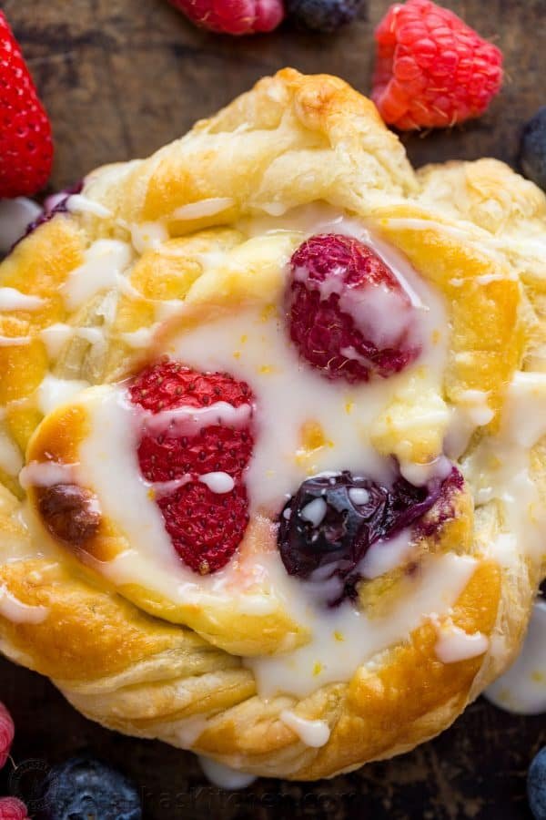 This Cream Cheese Danish recipe is so easy to make. The cheese danish filling tastes just like cheesecake and the fresh berries and lemon glaze add a bright pop of flavor. This puff pastry recipe always disappears fast! | natashaskitchen.com Up close cheese danish with lemon glaze and berries showing texture of puff pastry dessert recipe