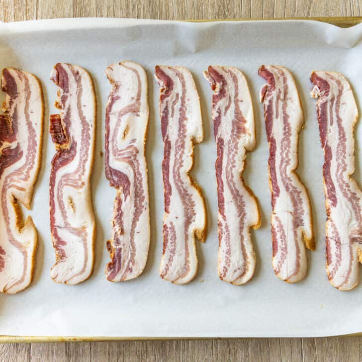 How to bake bacon in the oven that is perfectly crispy. Oven-baked bacon is the best for making a bigger batch and it's the easiest method. raw meat strips on a sheet pan ready for the oven