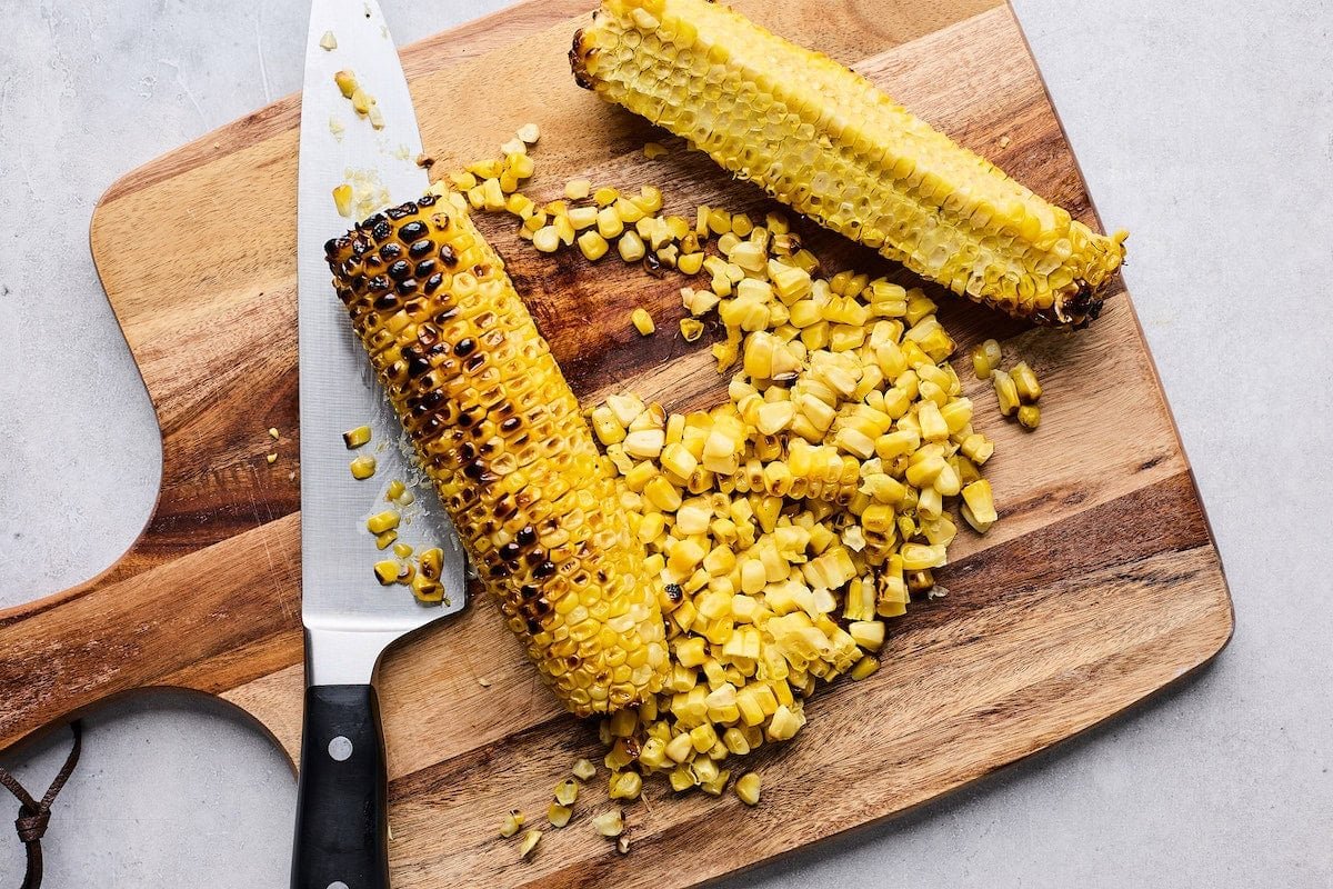 cutting charred corn off the cob with a sharp knife on wood cutting board.