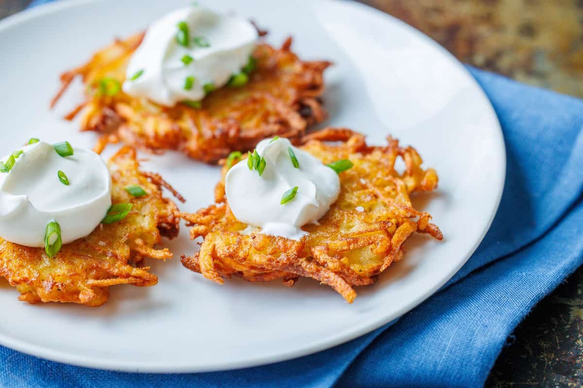 This latke recipe creates the crispiest potato fritters. Sprinkled with salt and dolloped with sour cream, these potato latkes are total comfort food. three latkes on a plate with a dollop of sour cream and sprinkled with fresh chives