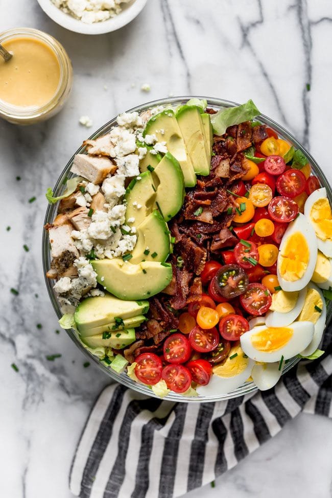 Cobb Salad With Chicken, Bacon, Tomatoes, Hard Boiled Eggs, Avocado, Blue Cheese, Chives, And A Simple Red Wine Vinegar Dressing. This Traditional Cobb Salad Is Loaded With Goodness And Always A Favorite! #salad #bacon #chicken #chickenrecipe #cobbsalad #avocado #eggs #glutenfree #glutenfreerecipe Cobb salad in large glass bowl with jar of dressing on the side and tea towel.