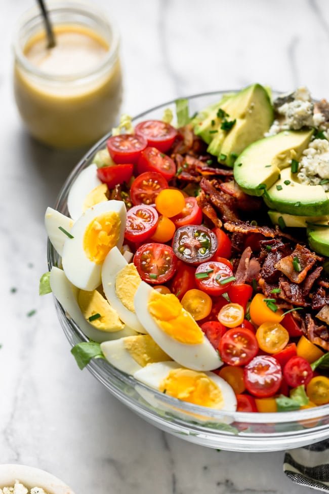 Cobb Salad With Chicken, Bacon, Tomatoes, Hard Boiled Eggs, Avocado, Blue Cheese, Chives, And A Simple Red Wine Vinegar Dressing. This Traditional Cobb Salad Is Loaded With Goodness And Always A Favorite! #salad #bacon #chicken #chickenrecipe #cobbsalad #avocado #eggs #glutenfree #glutenfreerecipe Cobb Salad in Bowl with salad dressing in jar