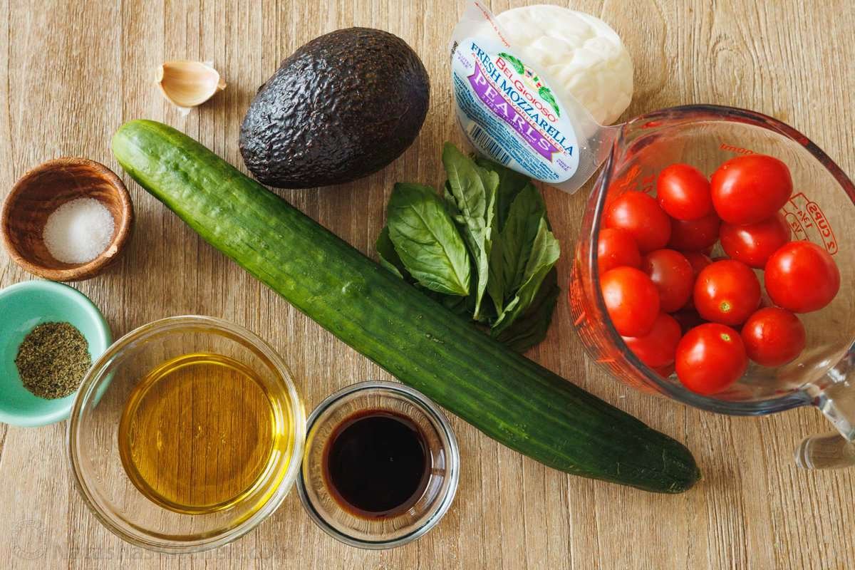 Ingredients for tomato and mozzarella salad including avocado, cucumber, basil leaves, cherry tomatoes, mozzarella pearls, garlic and balsalmic vinegar