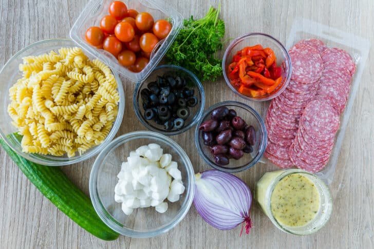 Ingredients for Italian pasta salad with rotini pasta, tomatoes, cucumber, olives, mozzarella, red onion, salami, parsley, roasted red pepper and homemade Italian Dressing