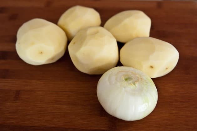 Five peeled potatoes and a peeled onion on a wooden cutting board 