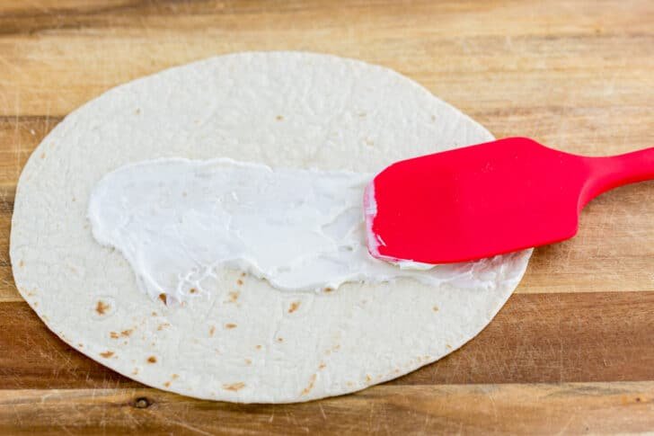 These Breakfast Burritos will make a breakfast person out of you. Crisp on the outside and filled with egg, ham, and cheese. Easy freezer breakfast burritos! A tortilla with a spatula spreading a dollop of sour cream