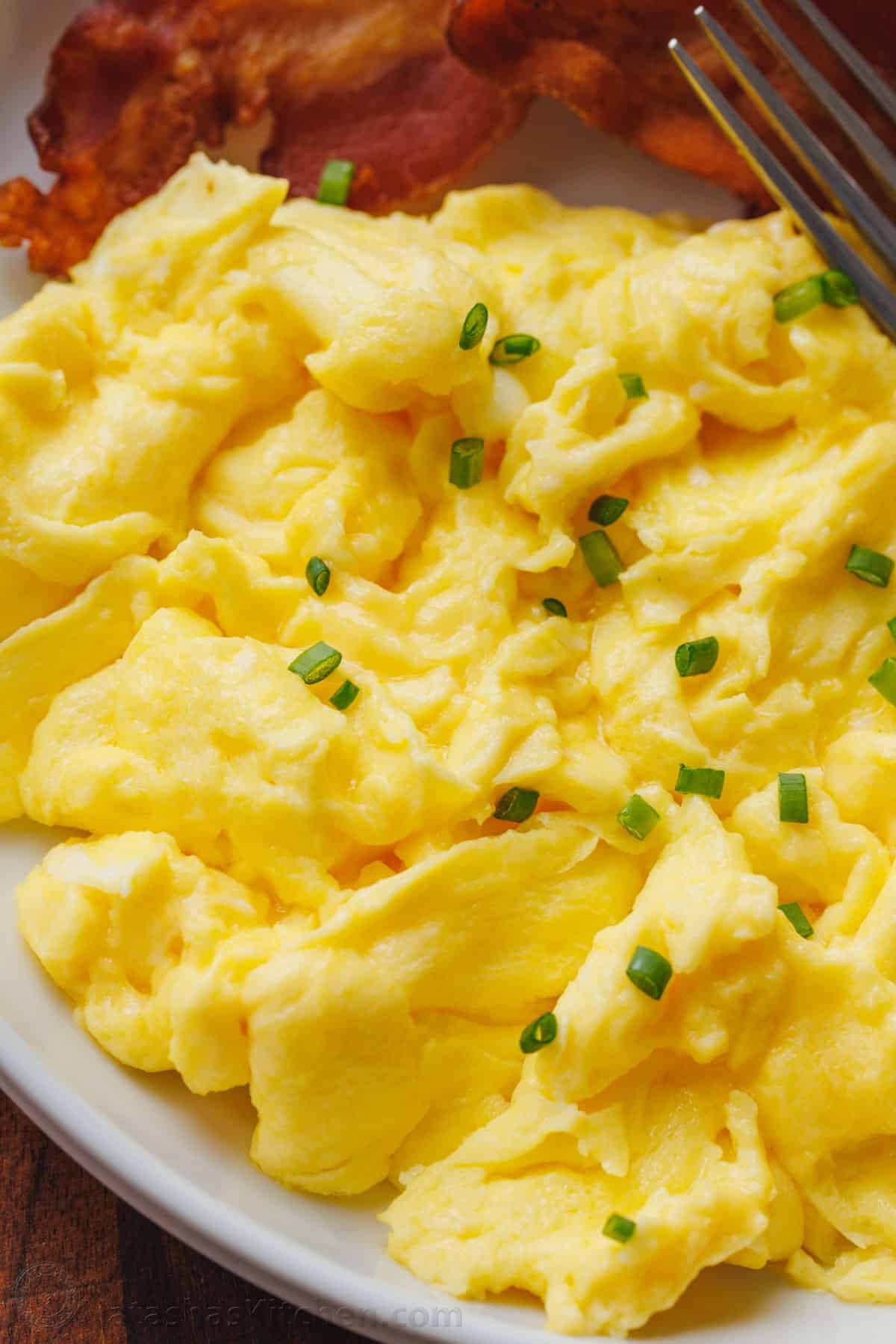 Learn how to cook perfect scrambled eggs that are soft, fluffy, and satisfying. This 5-minute recipe makes the absolute best scrambled eggs! Close up of fluffy scrambled eggs on a plate garnished with chives, next to crispy bacon.