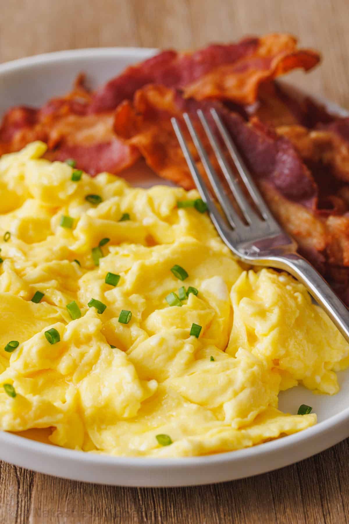 Learn how to cook perfect scrambled eggs that are soft, fluffy, and satisfying. This 5-minute recipe makes the absolute best scrambled eggs! Fluffy scrambled eggs on a plate garnished with chives, next to crispy bacon and a fork.