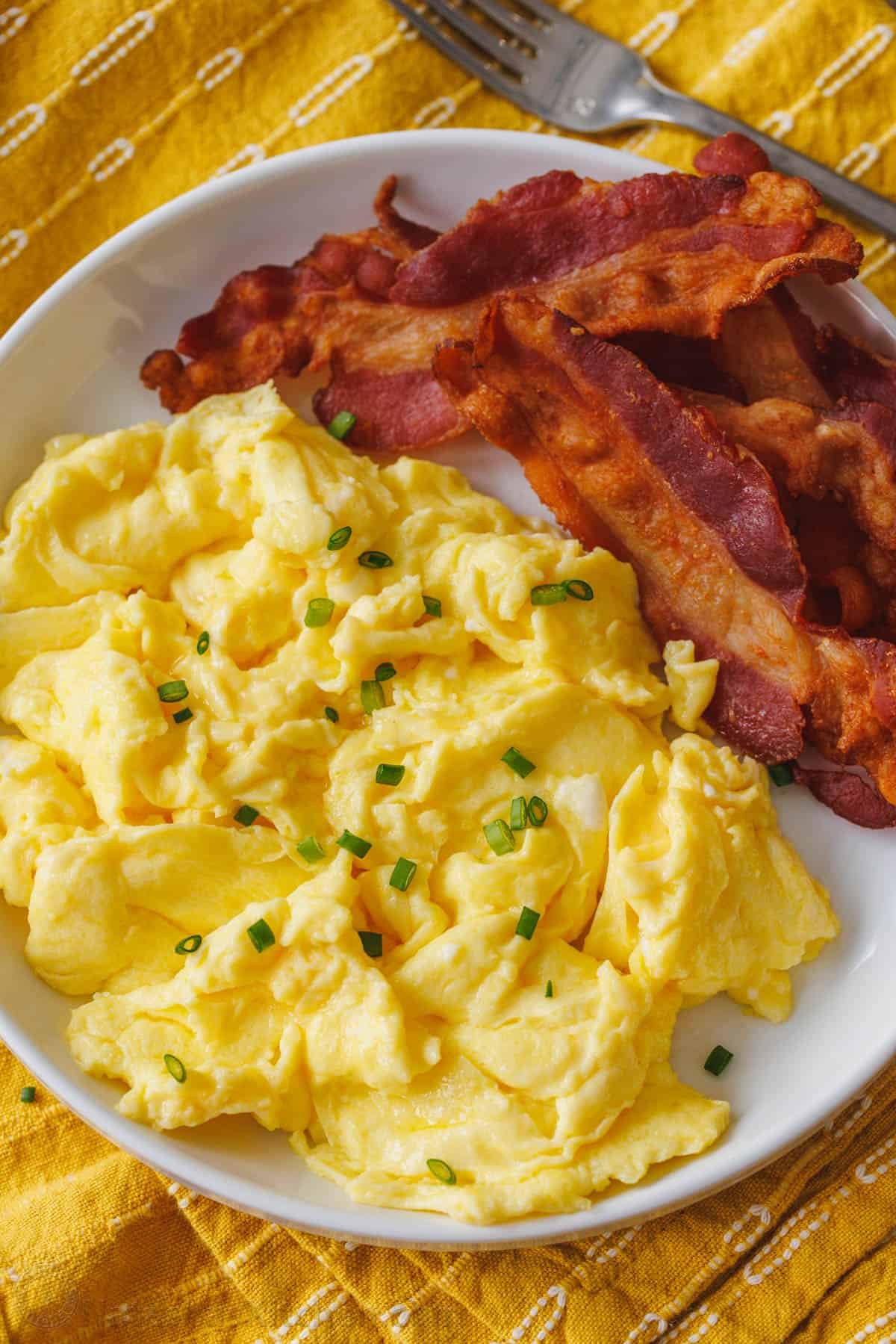 Learn how to cook perfect scrambled eggs that are soft, fluffy, and satisfying. This 5-minute recipe makes the absolute best scrambled eggs! Fluffy scrambled eggs on a plate garnished with chives, next to crispy bacon.