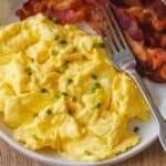 Fluffy scrambled eggs on a plate garnished with chives, next to crispy bacon and a fork.