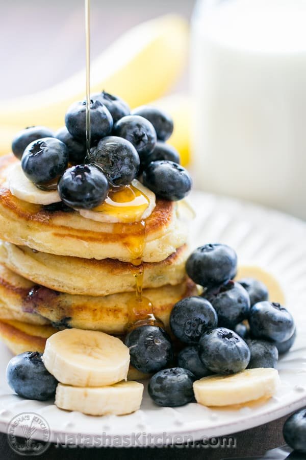 Blueberry pancakes stacked on a plate topped with syrup, blueberries, and bananas.