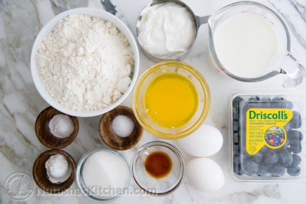 Pancake ingredients: flour, sugar, egg, milk, vanilla, salt, baking powder, baking soda, sour cream, blueberries in bowls/cups