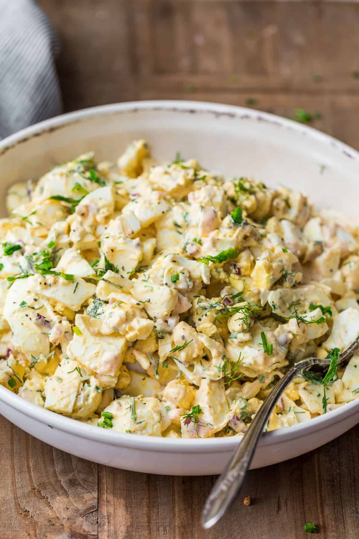 Our favorite Egg Salad Recipe with the best creamy dressing. Egg salad is an excellent side dish a great way to use leftover hard-boiled eggs. Quick Egg Salad in a serving bowl with a spoon and garnished with herbs,