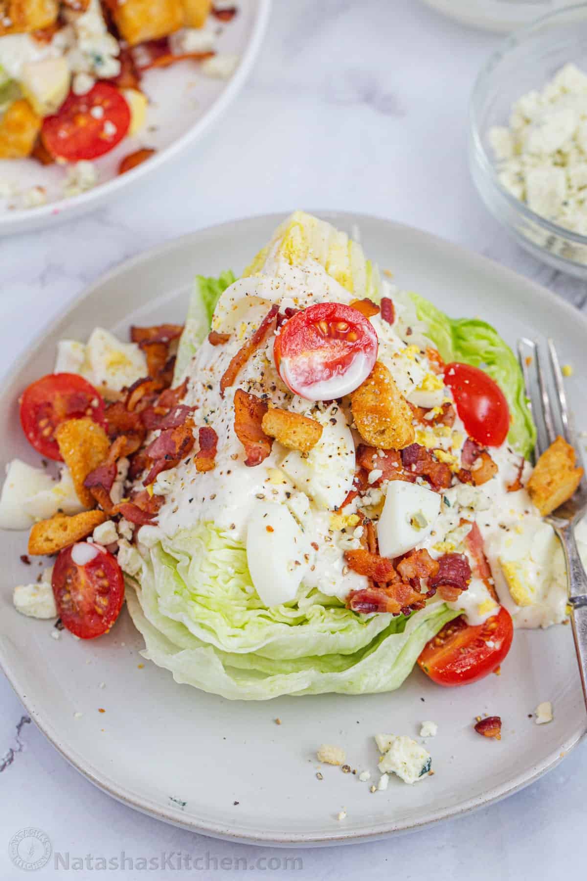 How to make the best Wedge Salad with iceberg lettuce, dressing, bacon, eggs, tomatoes, croutons, a creamy dressing, and blue cheese crumbles Closer up wedge of iceberg with dressing and toppings on a plate