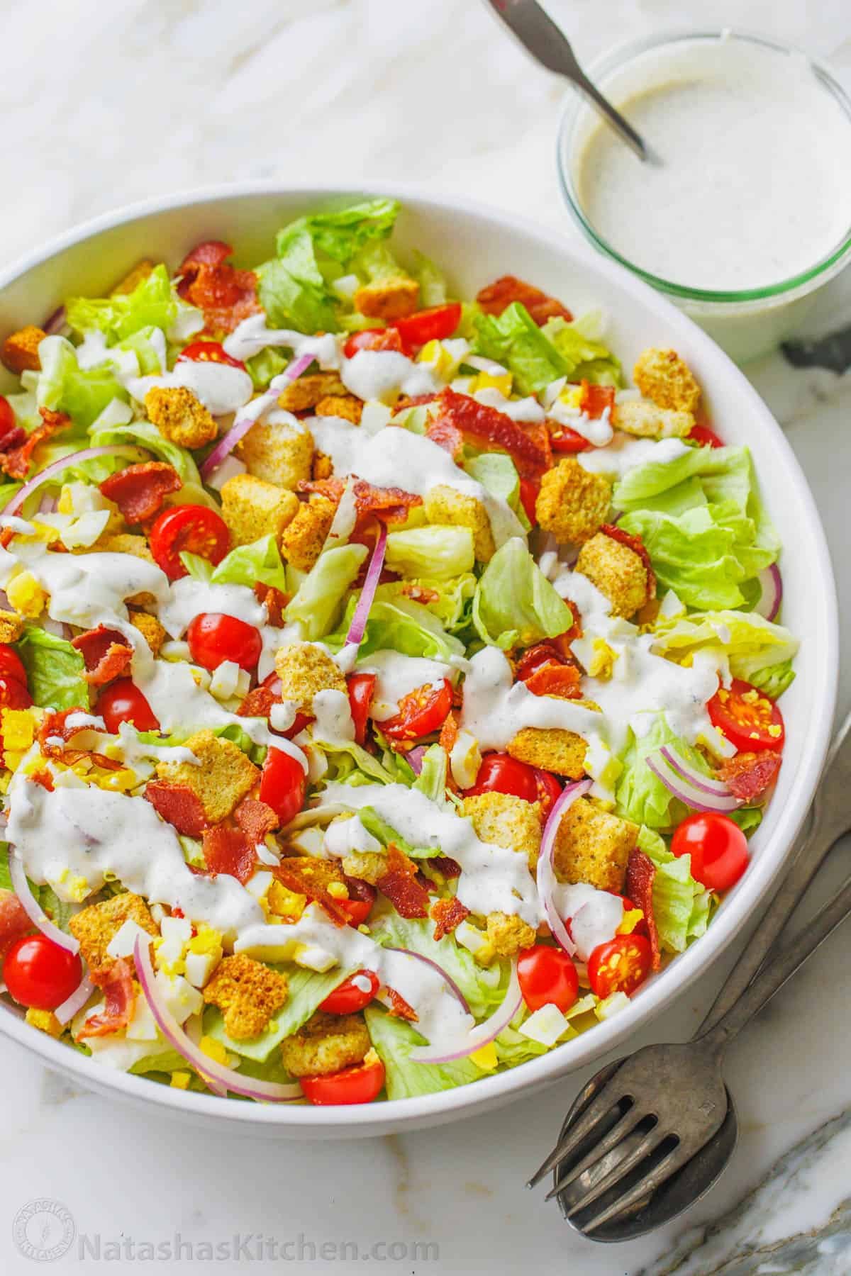 This restaurant-style Garden Salad is an easy salad recipe full of fresh ingredients and tossed with tangy homemade buttermilk dressing. A fresh garden salad in a large white bowl topped with croutons and drizzled with Ranch dressing, next to serving spoons and a bowl of dressing.