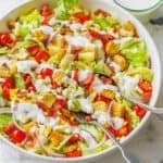 A fresh garden salad in a large white bowl topped with croutons and drizzled with Ranch dressing, with two serving spoons.
