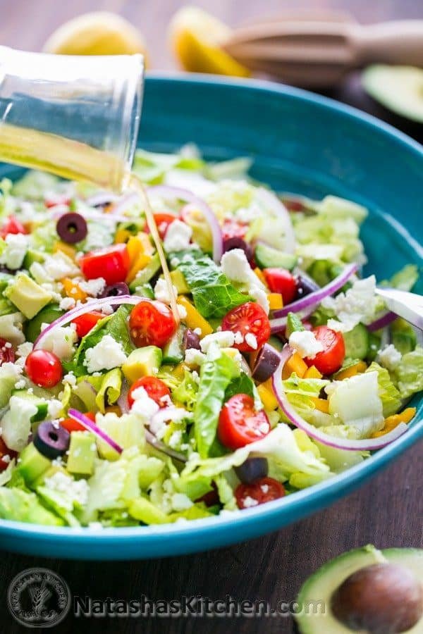 Fresh Greek Salad is a must try! So healthy with cucumber, tomato, avocado, olives and feta. You will want the easy Greek dressing on all of your salads! #greeksalad #greeksaladdressing #greekdressing #greekrecipes #salad #avocado #natashaskitchen Greek salad dressing pouring onto Greek salad