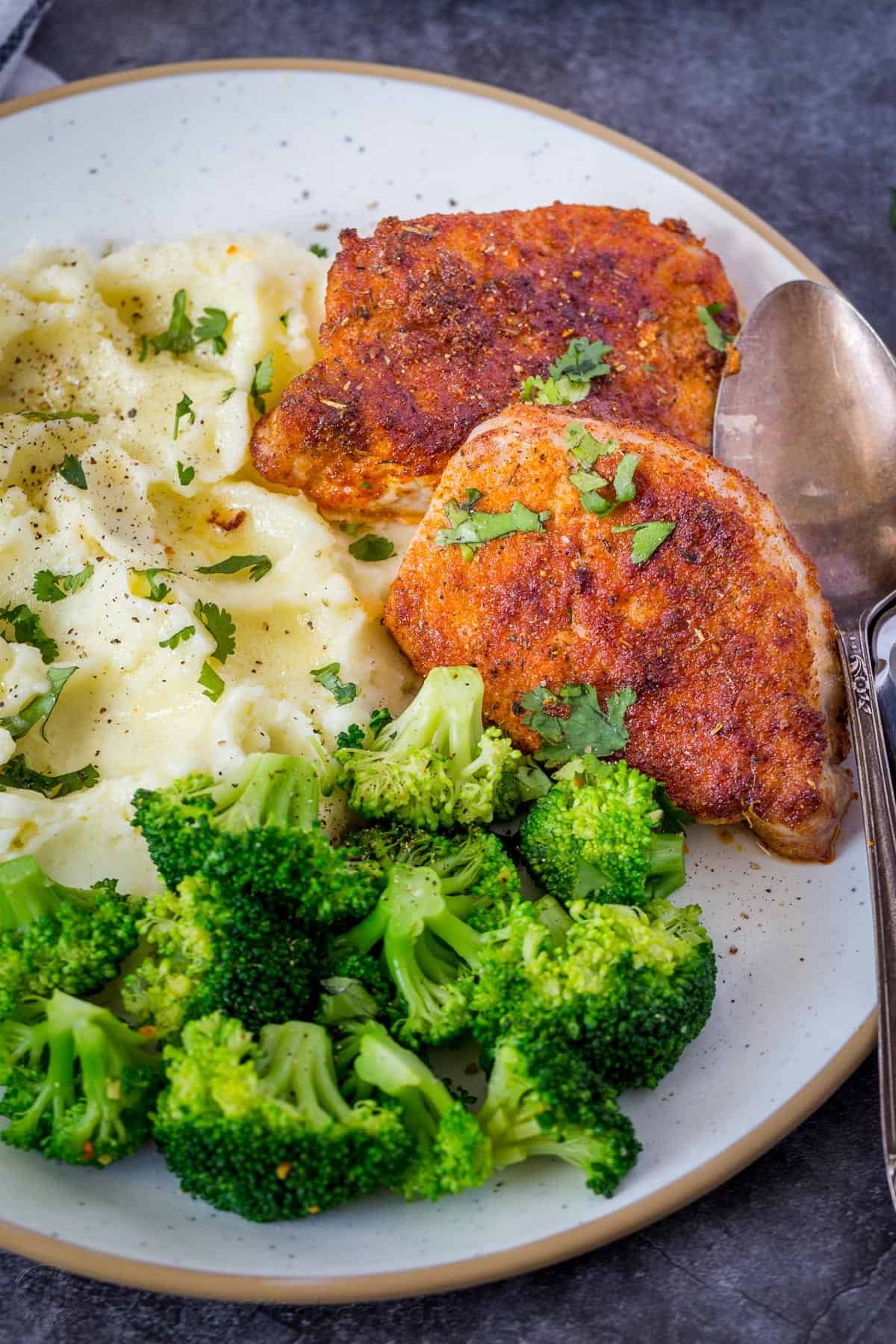 Seasoned pork chops in Air Fryer on a white plate with mashed potatoes and broccoli garnished with parsley.