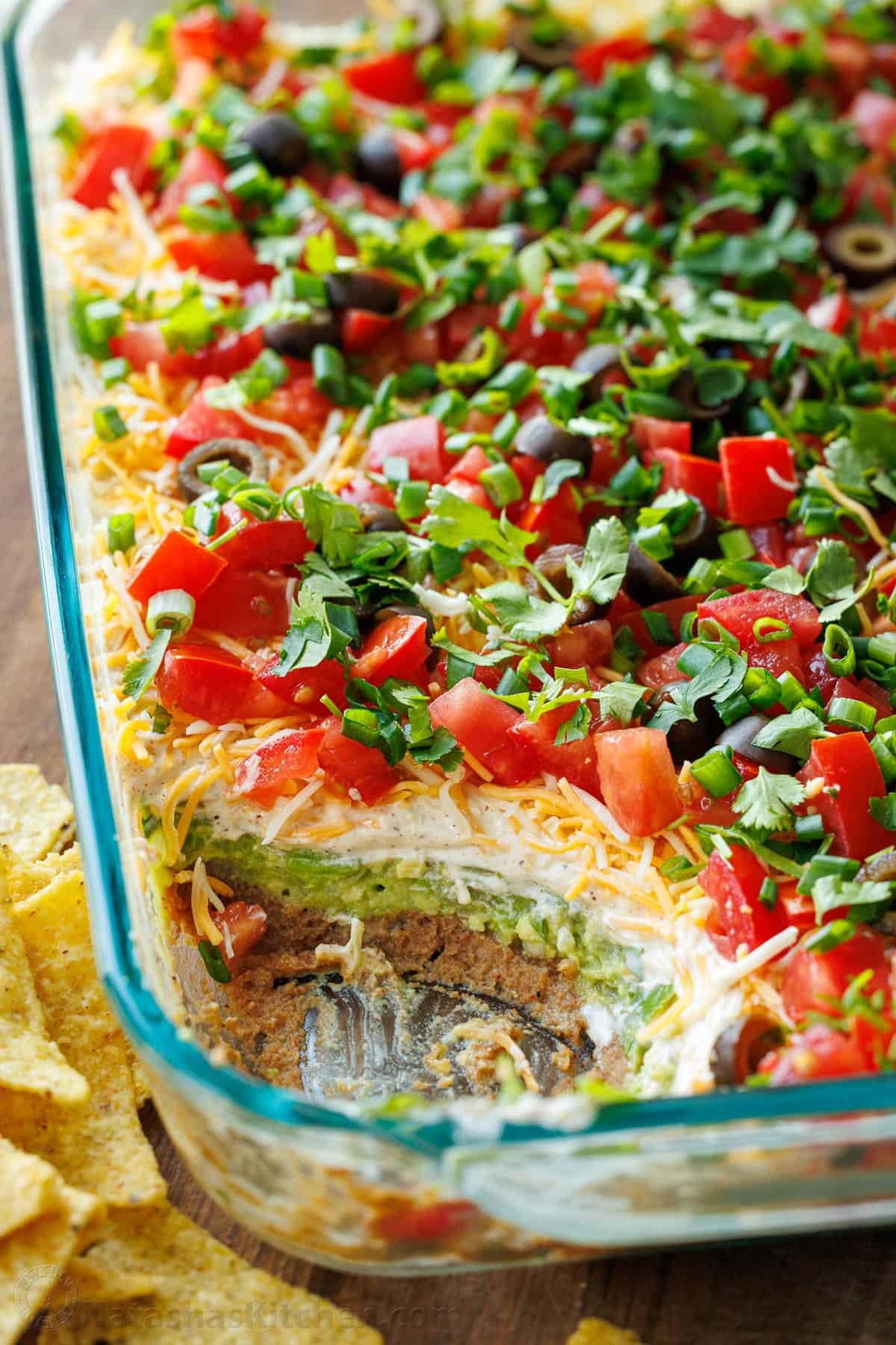 7-Layer Dip in a glass pan, showing each layer, refried beans, sour cream, cheese, tomatoes, green onion, guacamole, black olives with tortilla chips scattered around