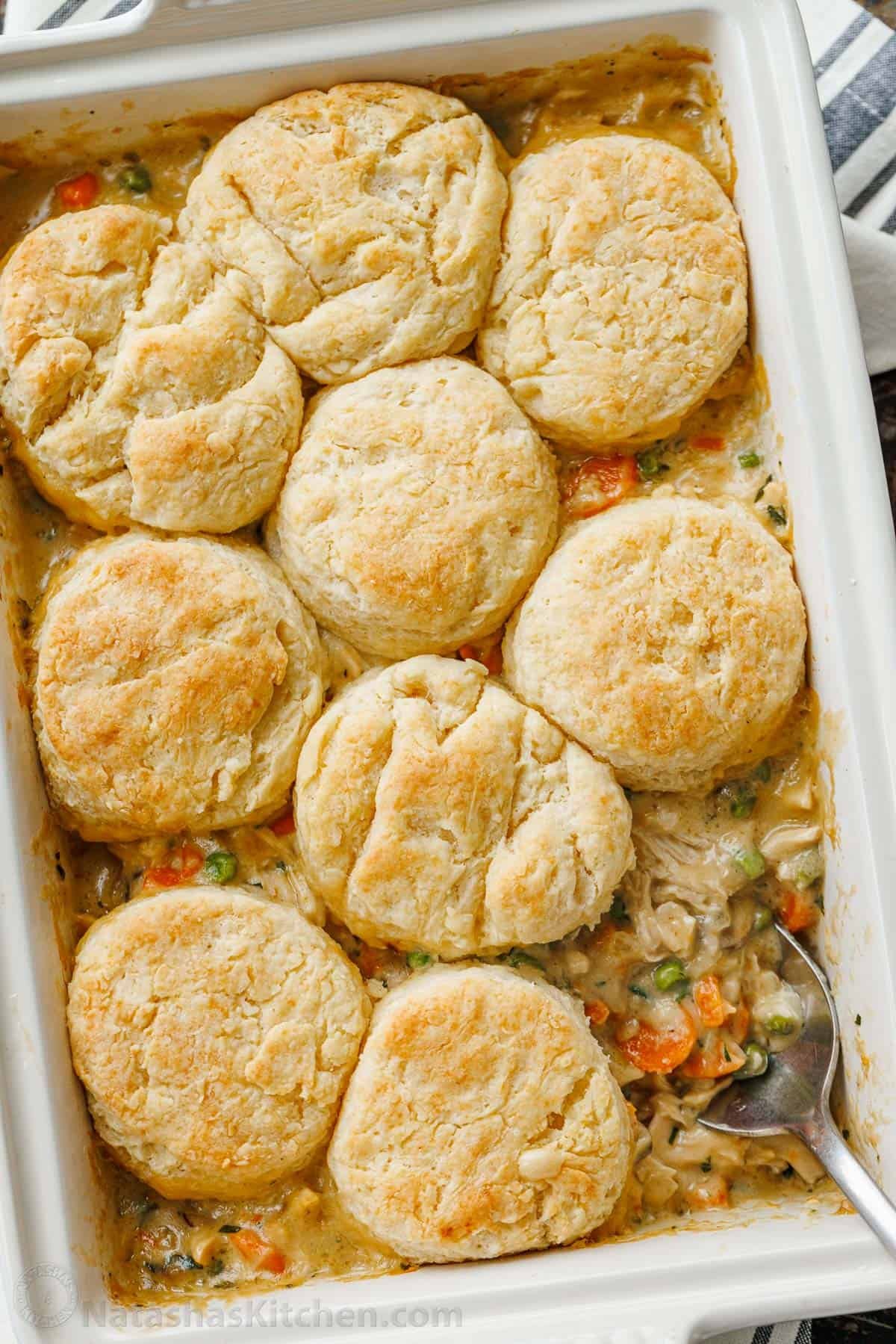 Chicken Pot Pie Casserole has all the cozy flavor of a creamy chicken pot pie, topped with flaky biscuits instead of baked inside a pie crust. Overhead view of a spoon scooping a serving of chicken pot pie casserole from a baking dish.