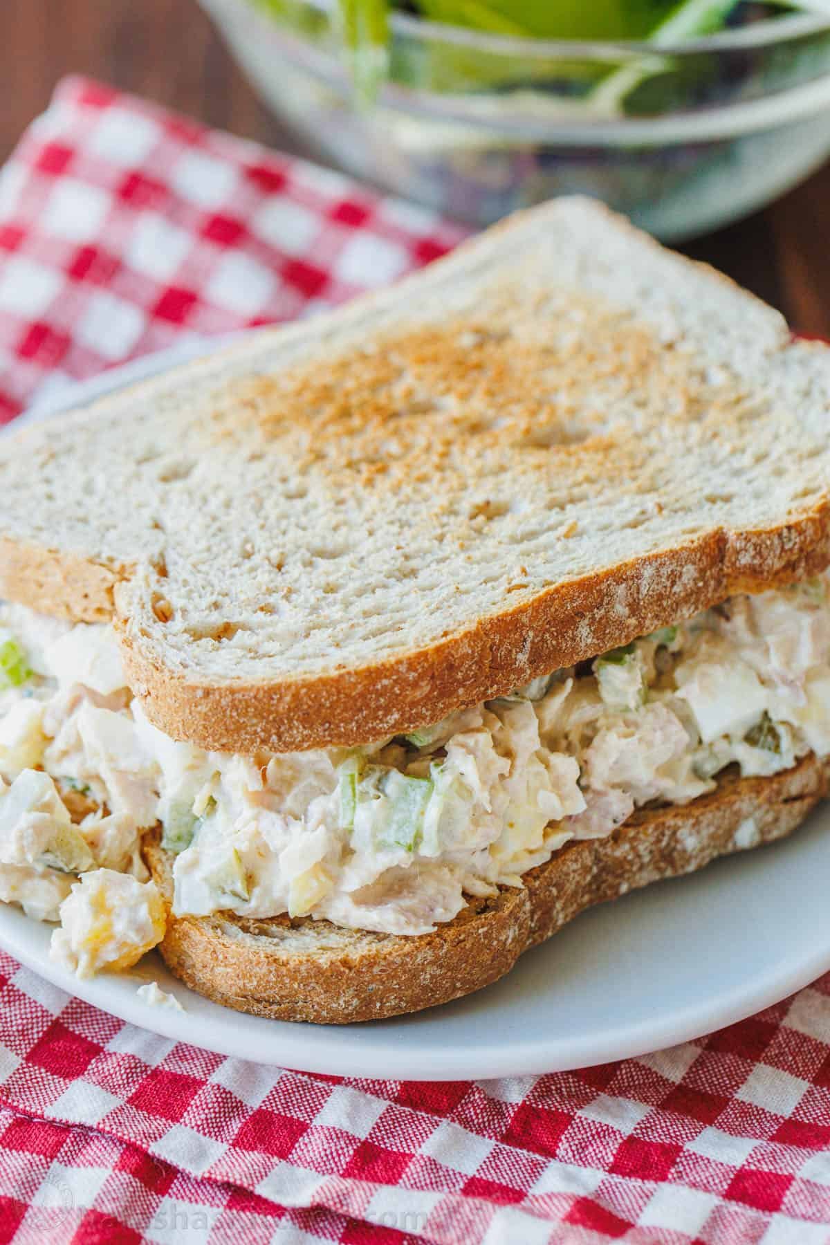 A classic tuna salad sandwich on a white plate, with a red and white checked cloth underneath.