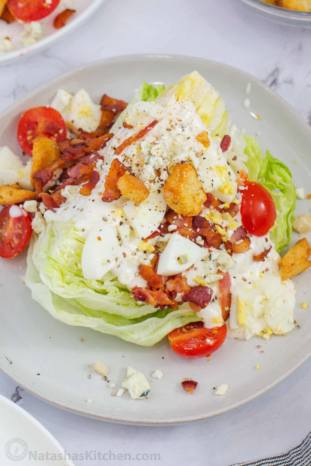 How to make the best Wedge Salad with iceberg lettuce, dressing, bacon, eggs, tomatoes, croutons, a creamy dressing, and blue cheese crumbles Lettuce wedge topped with dressing and toppings on a plate.