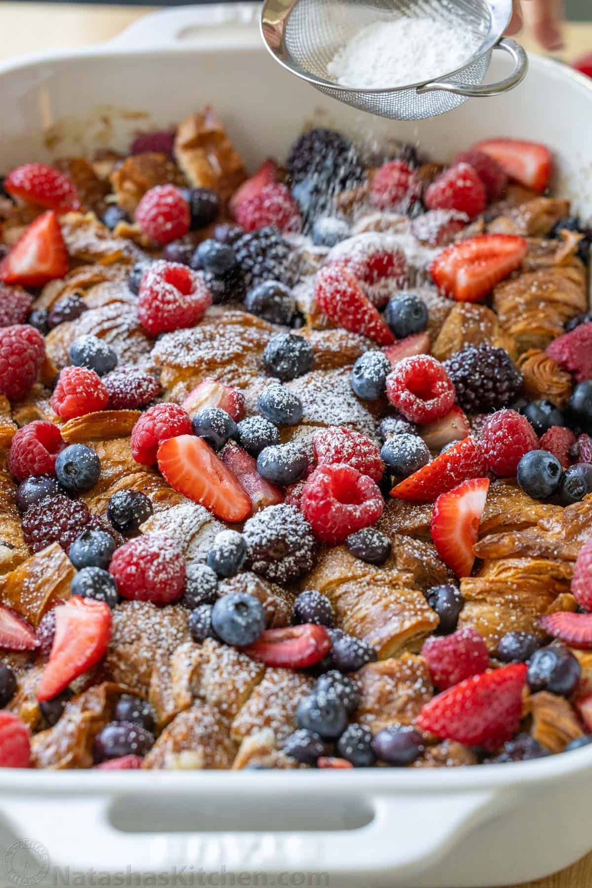 Dusting powdered sugar over croissant French Toast Casserole in a white dish