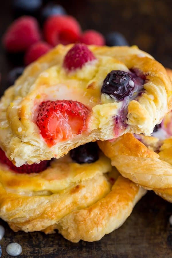 This Cream Cheese Danish recipe is so easy to make. The cheese danish filling tastes just like cheesecake and the fresh berries and lemon glaze add a bright pop of flavor. This puff pastry recipe always disappears fast! | natashaskitchen.com Stacked berry cheese danishes glazed with lemon glaze and showing the inside of the cheese danish puff pastry recipe