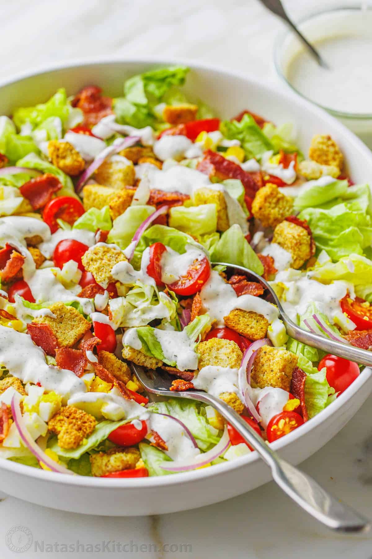 This restaurant-style Garden Salad is an easy salad recipe full of fresh ingredients and tossed with tangy homemade buttermilk dressing. Close up of homemade garden salad in a large white bowl topped with croutons and drizzled with Ranch dressing, with two serving spoons.