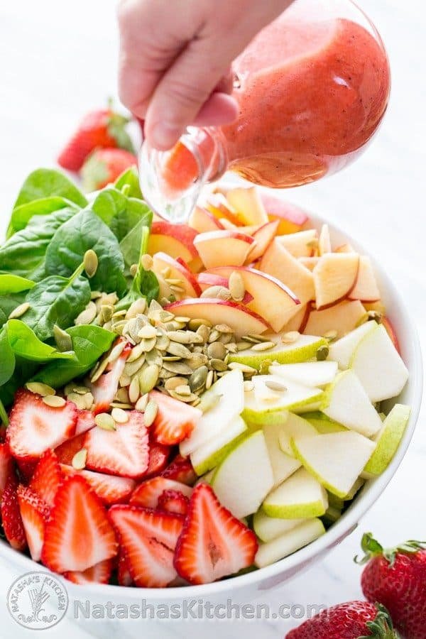 A fruit and spinach salad in a bowl with strawberry vinaigrette being poured on the salad 