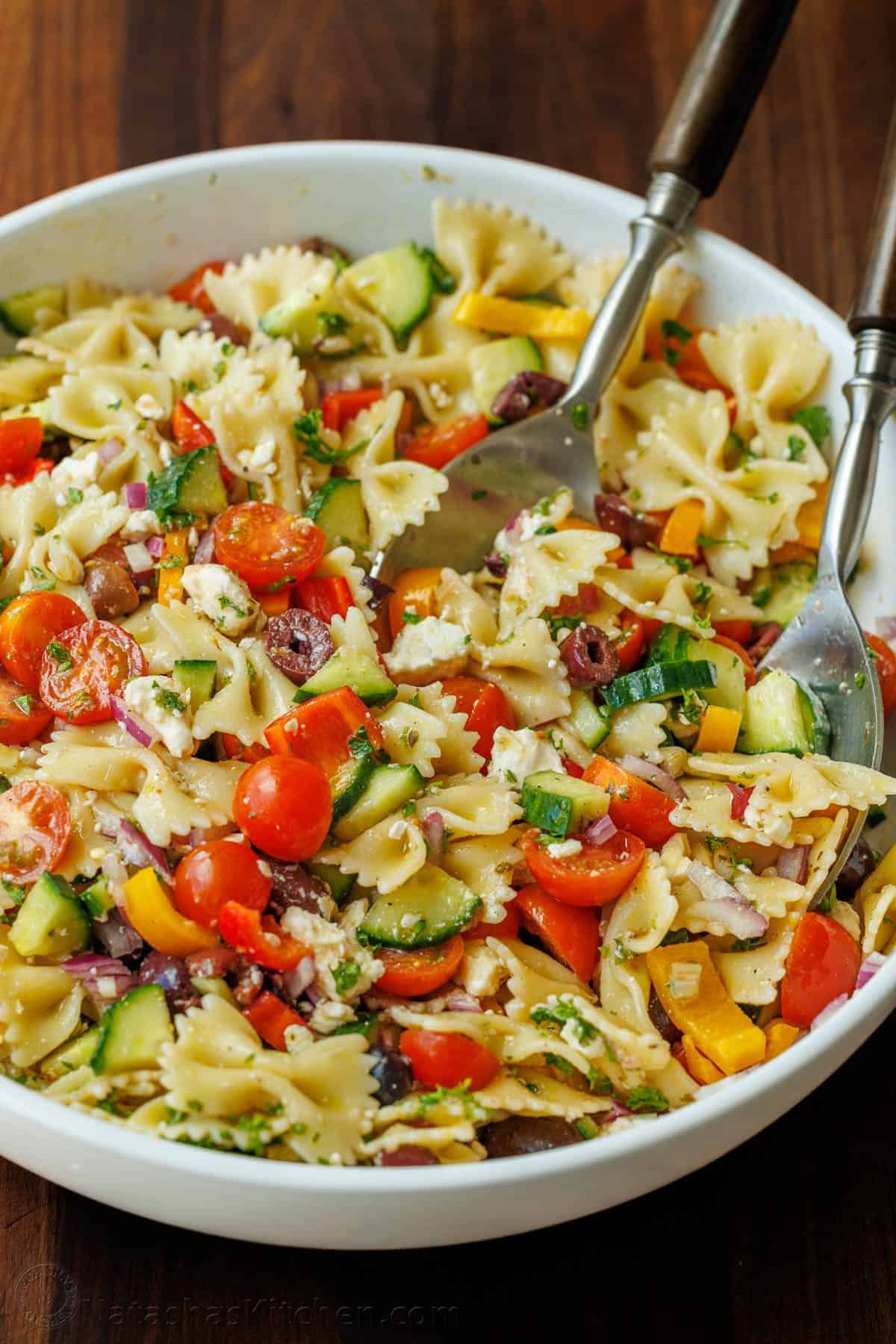 Greek Pasta Salad is a fresh, flavorful, and satisfying dish that brings a burst of Mediterranean flavor to any table. Perfect for any occasion and sure to please. Greek Pasta Salad ingredients tossed in a white serving bowl: bowtie pasta, cucumber, bell pepper, halved cherry tomatoes, Kalamata olives, red onion, feta cheese, and parsley