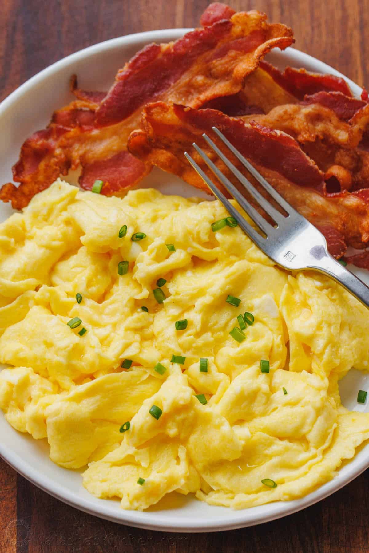 Learn how to cook perfect scrambled eggs that are soft, fluffy, and satisfying. This 5-minute recipe makes the absolute best scrambled eggs! Fluffy scrambled eggs on a plate garnished with chives, next to crispy bacon and a fork.