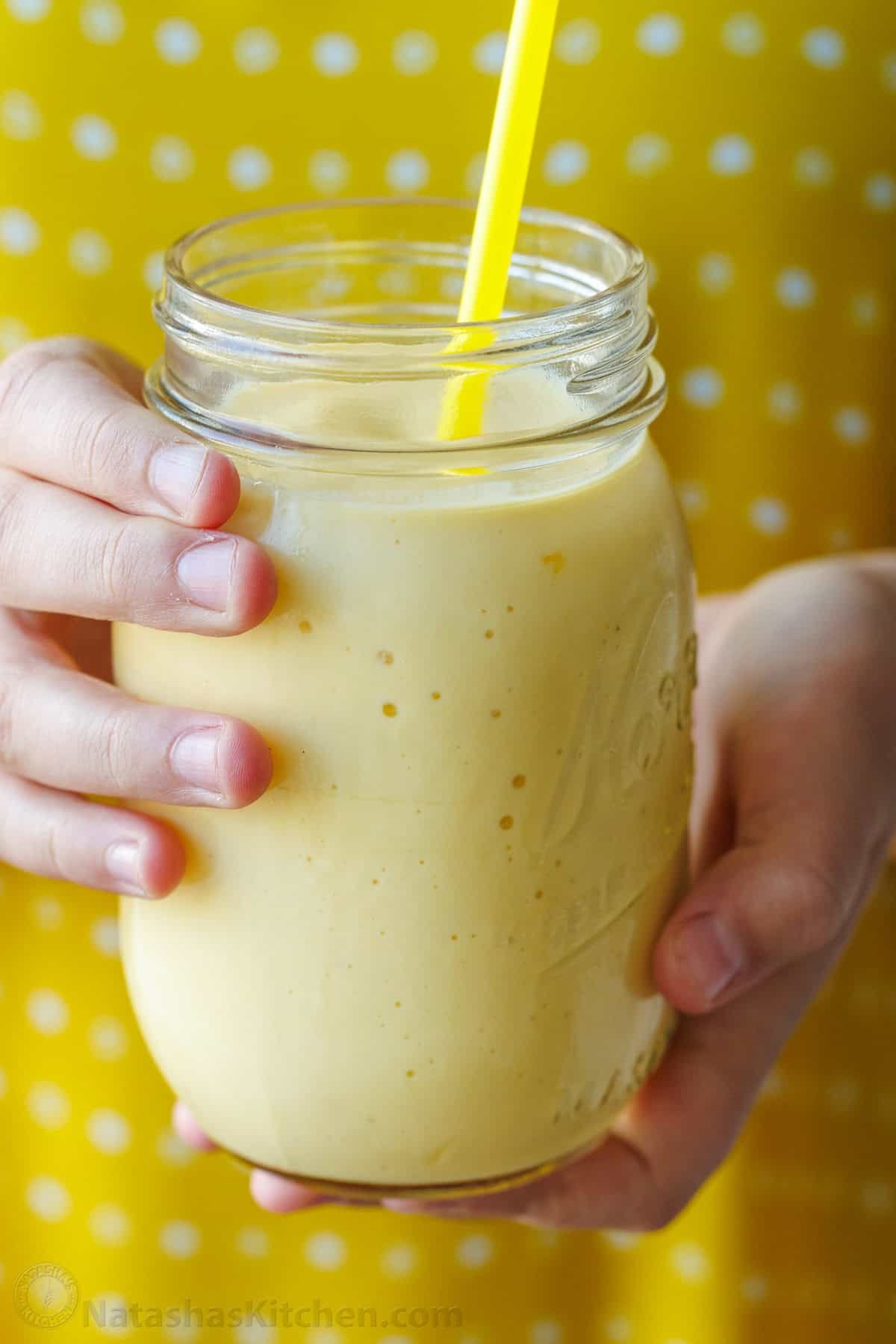 Easy Tropical Mango Smoothie recipe uses just 6 ingredients, like mango, lime, banana, and Greek yogurt, and takes under 5 minutes to make! hands holding a yellow mango smoothie in a mason jar with yellow polka-dot background