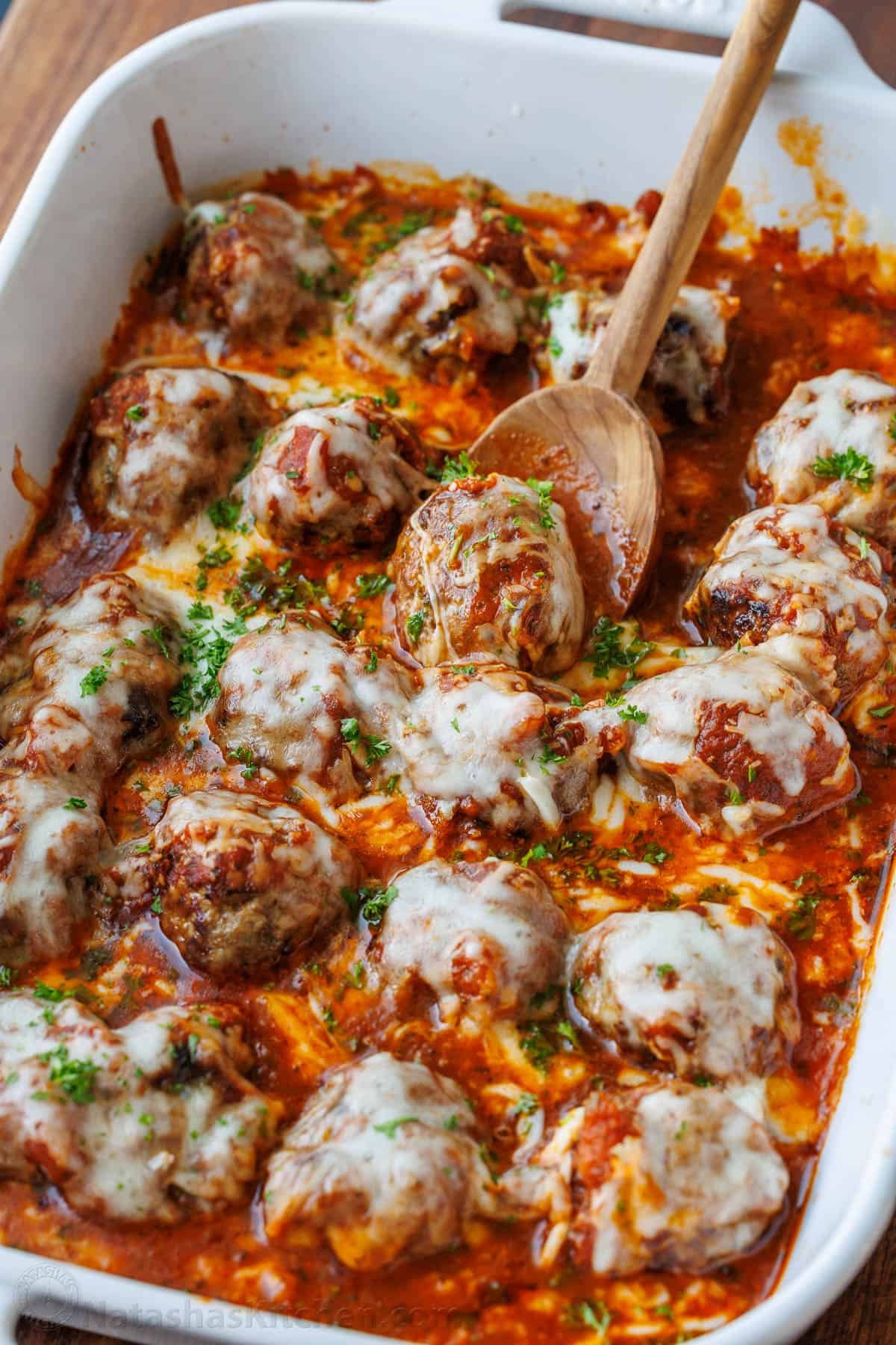 Meatballs Parmesan combines the best Italian dishes into one easy casserole. Homemade saucy Parmesan Meatballs topped with gooey cheese. Serving Italian meat ball casserole using a wooden spoon from a white casserole dish