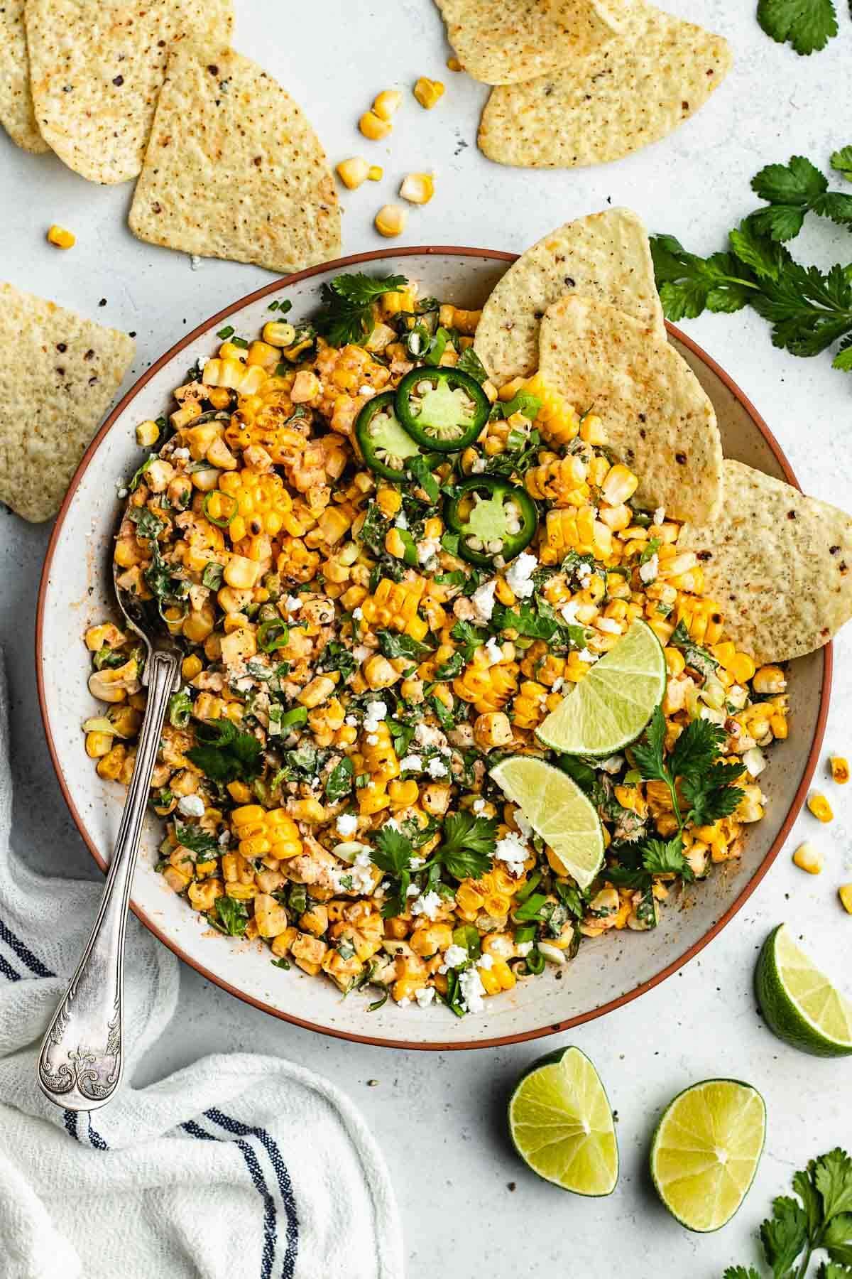 Mexican street corn salad in bowl with serving spoon, tortilla chips, and lime wedges.